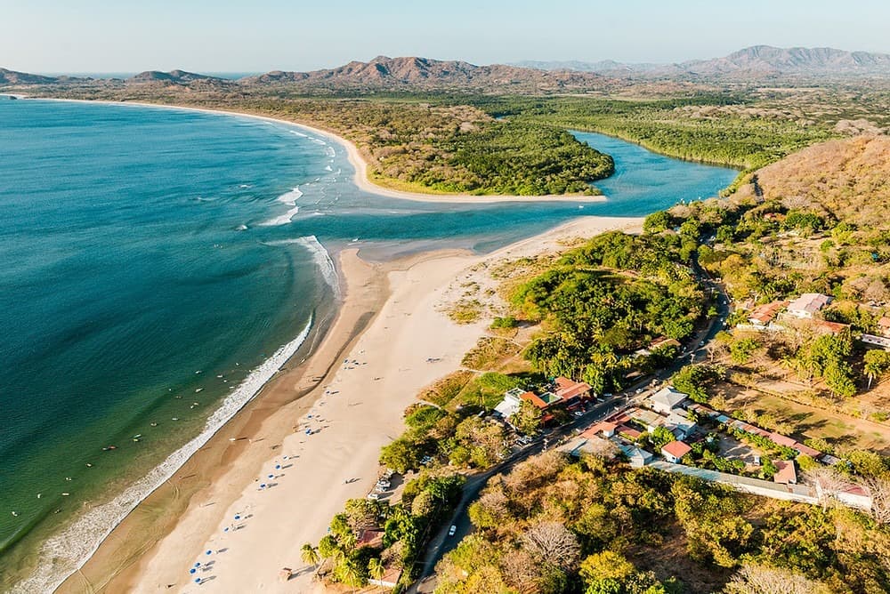 Aerial view of a long curving beach and river estuary on the Guanacaste coast, with the town and dry hills behind